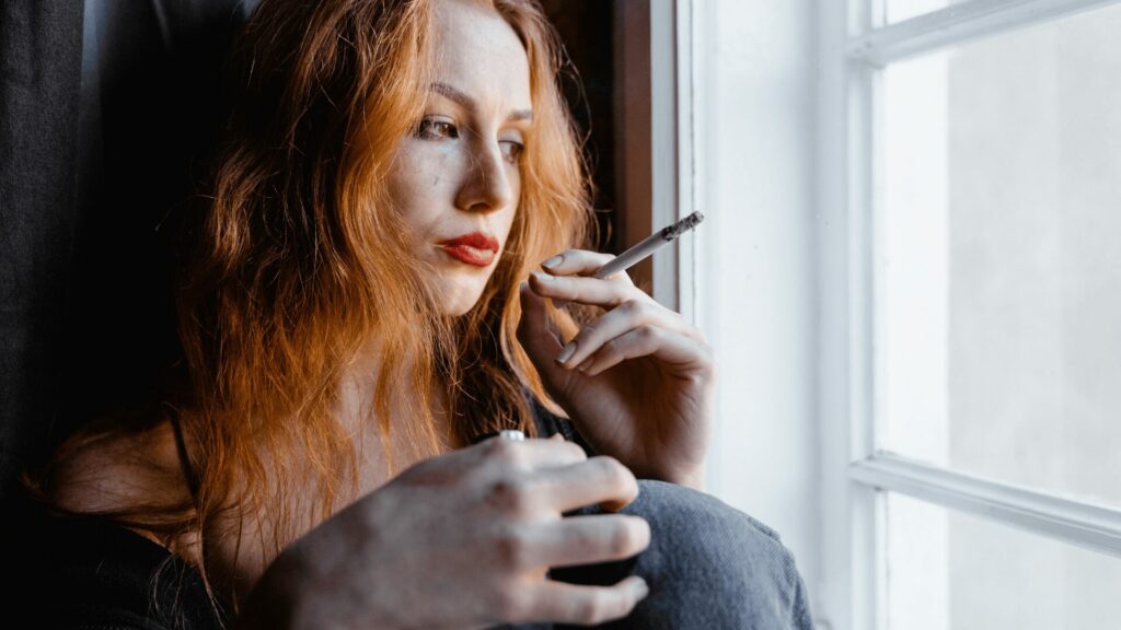 Femme songeuse fumant une cigarette devant une fenêtre, semblant réfléchir à l’arrêt du tabac et aux idées reçues, illustration des questionnements avant un accompagnement dans un centre LaserAddict.